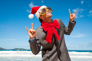 Funky businessman in Santa hat, sunglasses and big red Christmas bow throwing out peace signs on a tropical beach