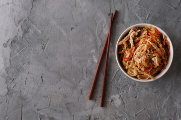 Fried udon noodles with chicken meat and sesame in white bowl on a gray concrete background. Flat lay, top view, copy space