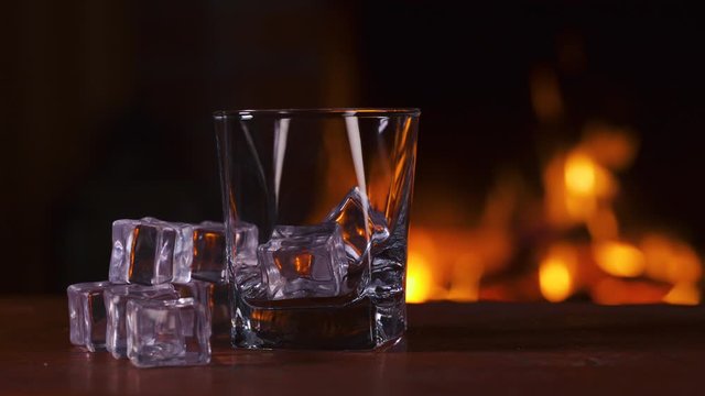 Empty glass with ice stands on a wooden table against the background of a fireplace with flame. Alcoholic drink concept. Fire background. Drink alone. Macro. Close up.