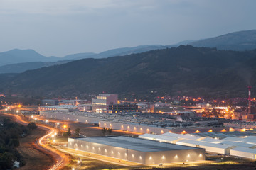 Close up view from above of a large, modern, illuminated factory and curvy road with car trails