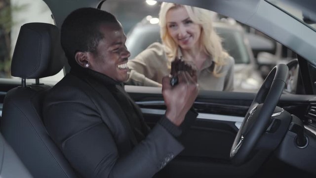 Blurred Caucasian woman giving car keys to the African American man sitting in salon. Satisfied customer gesturing yes and smiling. Successful female car dealer selling automobile in showroom.