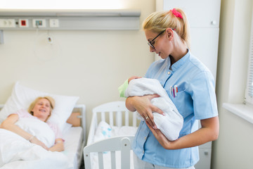 Young nurse standing in maternity ward and holding newborn baby in her arms. After birth concept.