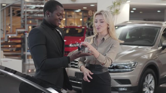 Pretty Caucasian woman giving car keys to the African American man in automobile showroom. Happy client gesturing yes and smiling, saleswoman clapping hands. Car dealership, car business.