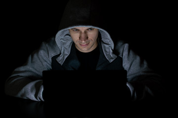 A man in a hood sits in the dark in front of a laptop screen. Spy, cybercriminal close up