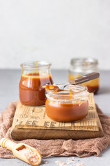 Homemade salted caramel sauce in glass jars on wooden cutting board. Selective focus.