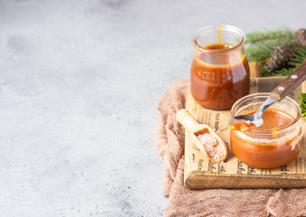 Spoon with tasty caramel sauce over glass jar as a gift for Christmas or New year with fir branches and cones. Selective focus.