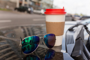White paper Cup of coffee with the red cover on the hood of the car. Paper Cup with hot tea on the hood of the SUV, closeup on the background of the windshield