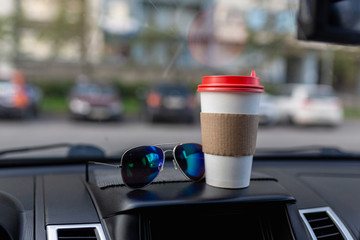 White paper coffee Cup and sunglasses on the dashboard of the car. Paper Cup with hot tea and glasses on the dashboard of the SUV close up against the background of a blurred Parking lot
