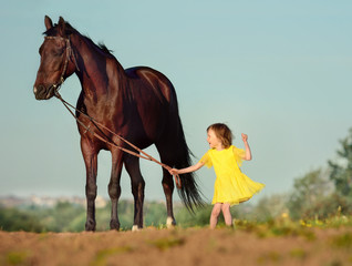 Fototapeta premium Happy smiling young girl five years old, in yellow dress holding in little hand the reins and big horse.