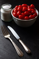 CHERRY RED TOMATOES IN BOL NEXT TO BOAT WITH SALT, FORK AND KNIFE ON BLACK BACKGROUND