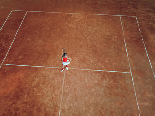 A young woman with a tennis racket in her hand stands in the center of a tennis court and looks into the camera. Top view, aerial view