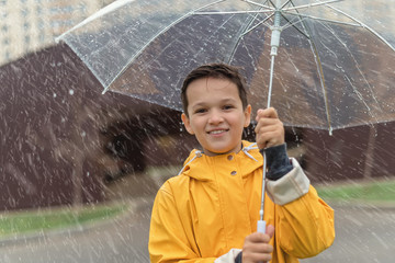 Handsome smiling brunette boy in yellow raincoat under transparent umbrella at rainy day