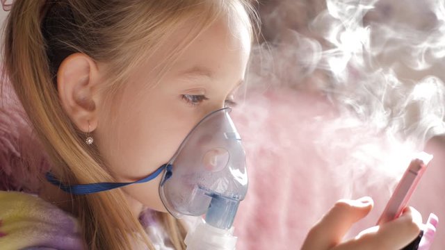 Little girl holds inhaler mask at home. Sick kid breathes through a nebulizer. Baby using equipment to treat asthma or bronchitis. Home treatment.