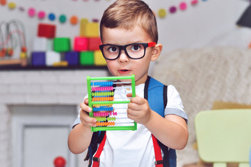 A little boy with a backpack on his back holds an abacus in his hands.