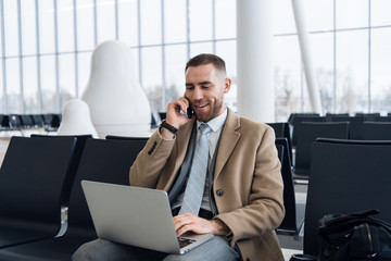 Happy businessman working on the laptop and talking on cellphone at the airport waiting lounge. Handsome caucasian businessman at waiting room in airport terminal