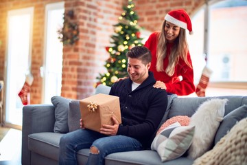 Young beautiful couple smiling happy and confident. Woman surprise man with gift  around christmas tree at home