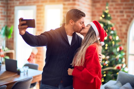 Young beautiful couple smiling happy and confident. Hugging and kissing make selfie by camera standing around christmas tree at home