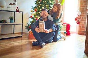 Obraz na płótnie Canvas Young beautiful couple smiling happy and confident. Woman covering eyes of man surpise her with gift around christmas tree at home