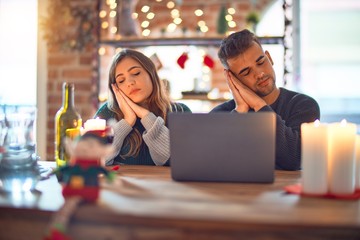 Young beautiful couple sitting using laptop around christmas decoration at home sleeping tired dreaming and posing with hands together while smiling with closed eyes.
