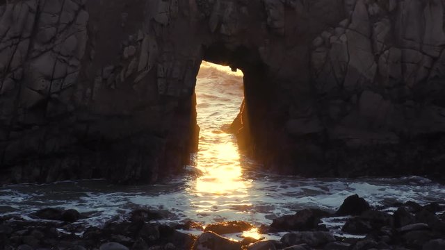Zoom out shot of sunlight glistening on the ocean waves through the framing of a naturally arched keyhole sea stack rock at sunset in Big Sur California.