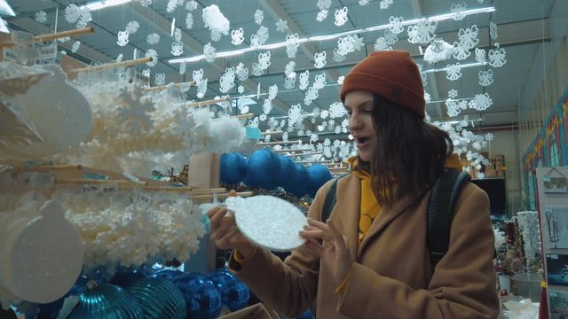 Beautiful hipster girl in a yellow tracksuit and a brown coat with an orange hat chooses Christmas and New Year decorations in a store and walks between shelving. Having fun. Slow motion. High resolut
