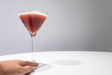 Cocktail glass and woman's left hand touching base of glass with red colored cocktail on white table gray background. Drink concept. Sangria cocktail. Selective focus. Copy space.