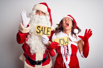 Couple wearing Santa costume holding wow and sale banner over isolated white background crazy and mad shouting and yelling with aggressive expression and arms raised. Frustration concept.