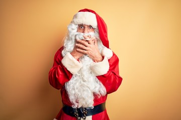 Middle age handsome man wearing Santa costume standing over isolated yellow background shocked covering mouth with hands for mistake. Secret concept.