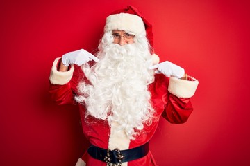 Obraz na płótnie Canvas Middle age handsome man wearing Santa costume standing over isolated red background looking confident with smile on face, pointing oneself with fingers proud and happy.