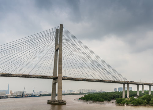 Ho Chi Minh City, Vietnam - March 12, 2019: Long Tau and song Sai Gon rivers meeting point. Landscape with H-shaped pylon of Phu My suspension bridge in center under gray cloudscape. Brown water. © Klodien