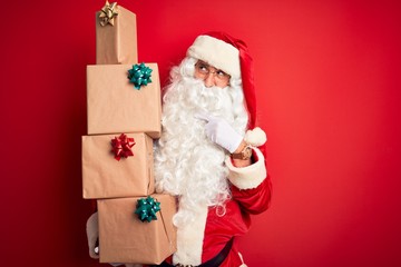 Senior man wearing Santa Claus costume holding tower of gifts over isolated red background serious...