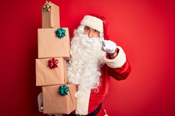 Senior man wearing Santa Claus costume holding tower of gifts over isolated red background pointing with finger to the camera and to you, hand sign, positive and confident gesture from the front