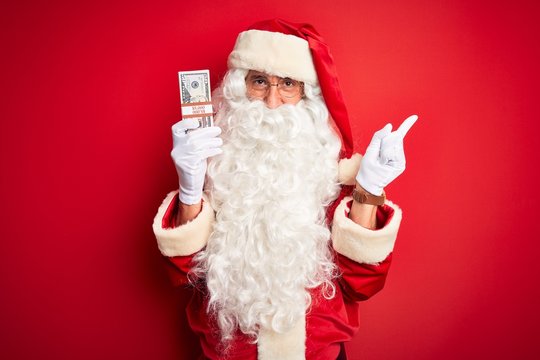 Middle age man wearing Santa Claus costume holding dollars over isolated red background very happy pointing with hand and finger to the side
