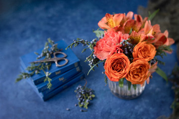 Vase of orange roses, carnations, and alstroemeria in clear vase on blue textured background; vintage scissors on small blue books in background; sprigs of blue cedar berries surround vase.