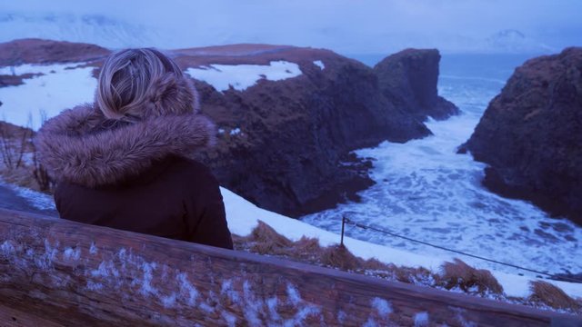 Iceland Female Sitting On Bench Looking At Cliffs And The Ocean In Arnarstapi In Winter 3
