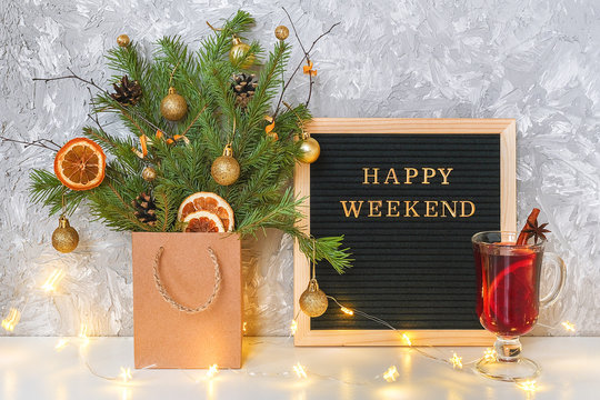 Happy Weekend text on black letter board, festive decored christmas tree in craft package, light lamp garland and glass of hot mulled wine with spices on table. Concept Hello winter weekend