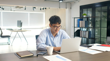 Young asian businessman smiling while working with laptop computer at office, business office lifestyle concept