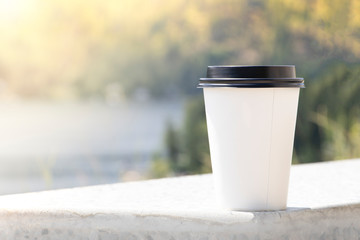 A paper cup of take away coffee on bench with blurry nature and sun background. Concept of start every morning with some drink make you feel fresh. Free copy space for text on left