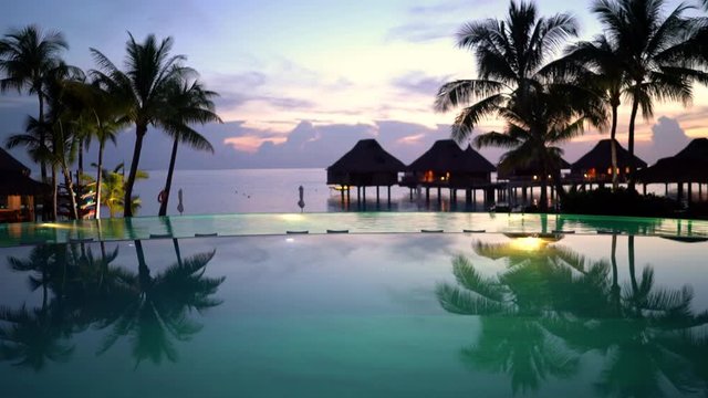 SEAMLESS LOOP VIDEO: Pool vacation travel destination - beach, palm trees and overwater bunglow hotel resort and luxury infinity swimming pool at amazing tropical sunset.