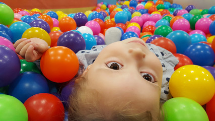 Fototapeta na wymiar Happy cute girl having fun in ball pit in kids amusement park and indoor play center. Close up portrait child playing with colorful balls in playground ball pool. Activity toys for little kid.