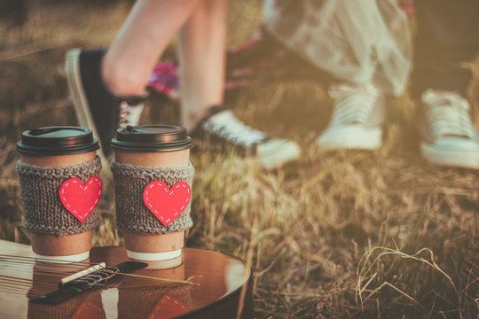 Romantic Couple Hugging While Having Picnic In Sunset Light. Coffee Cup Cozy Knitted Sleeve With Felt Red Heart.