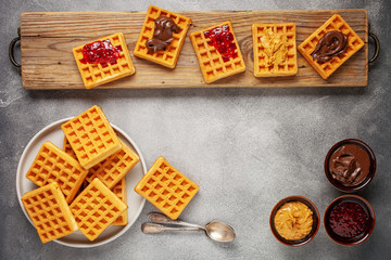 Belgian waffles with raspberry jam, chocolate and peanut paste in a cutting board on a grey background. Top view. Flat lay