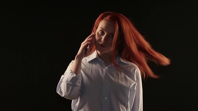 beautiful red-haired girl in a white shirt talking on a smartphone isolated on a black background. Phone advertising. Businesswoman in work