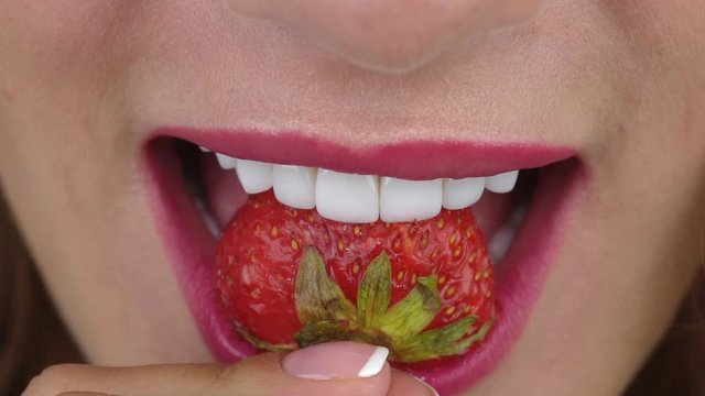 girl with dental veneers on white straight teeth and pink lipstick bites big juicy strawberry slow motion extreme closeup