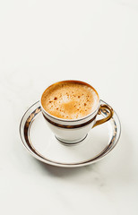 Cup of fresh americano or espresso coffee with golden foam froth on pile of brown raw coffee beans on white marble table background. Morning hot drink, coffee break, cope space