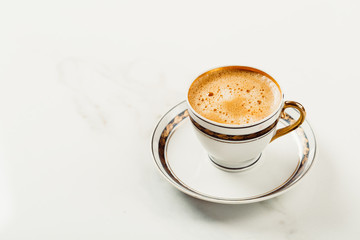 Cup of fresh americano or espresso coffee with golden foam froth on pile of brown raw coffee beans on white marble table background. Morning hot drink, coffee break, cope space