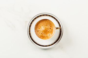 Cup of fresh americano or espresso coffee with golden foam froth on pile of brown raw coffee beans on white marble table background. Morning hot drink, coffee break, cope space