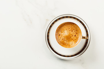 Cup of fresh americano or espresso coffee with golden foam froth on pile of brown raw coffee beans on white marble table background. Morning hot drink, coffee break, cope space
