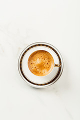 Cup of fresh americano or espresso coffee with golden foam froth on pile of brown raw coffee beans on white marble table background. Morning hot drink, coffee break, cope space