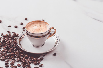 Cup of fresh americano or espresso coffee with golden foam froth on pile of brown raw coffee beans on white marble table background. Morning hot drink, coffee break, cope space